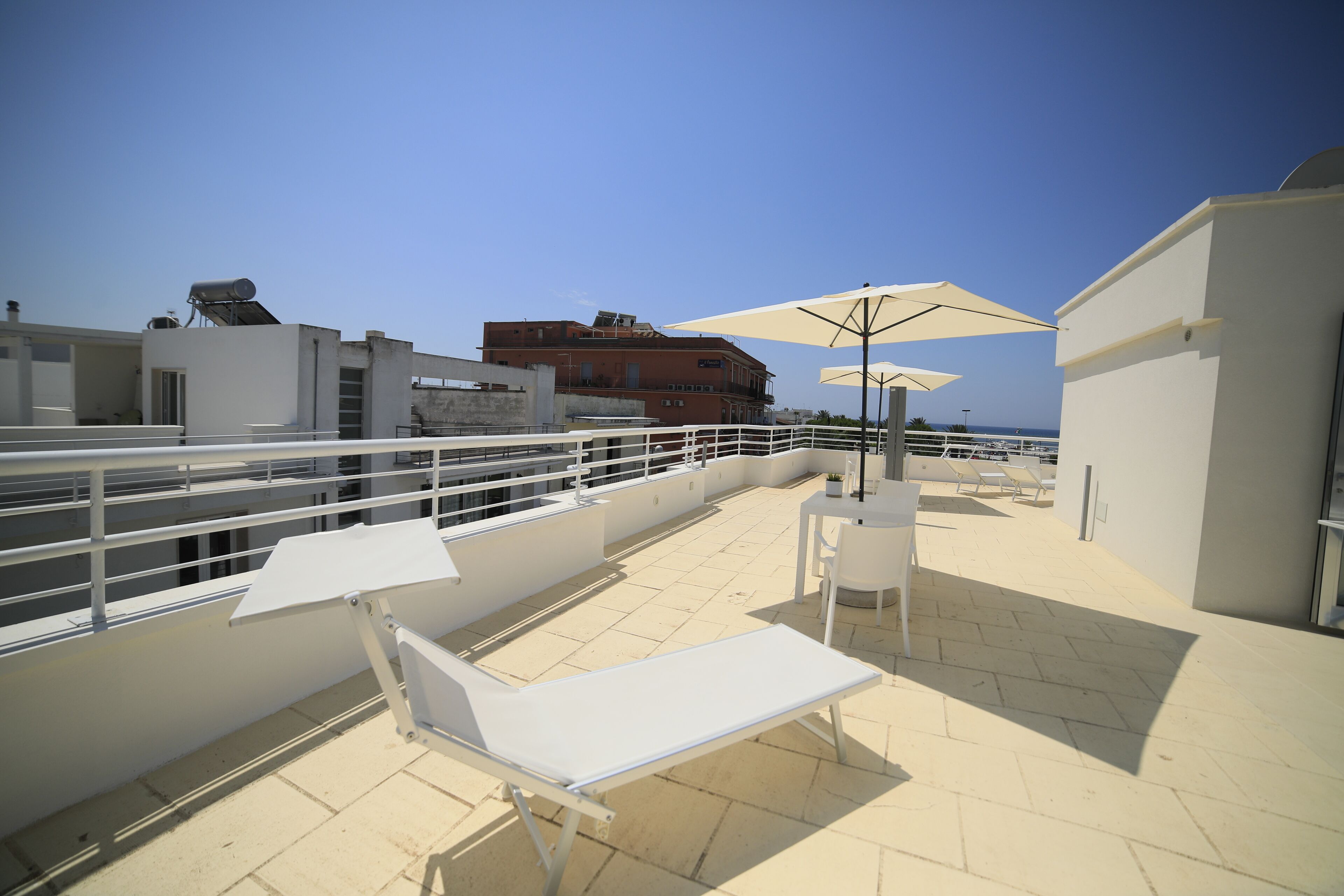 Eine Dachterrasse mit weißen Liegestühlen und Sonnenschirmen, einem Geländer und Blick auf Gebäude sowie das Meer.