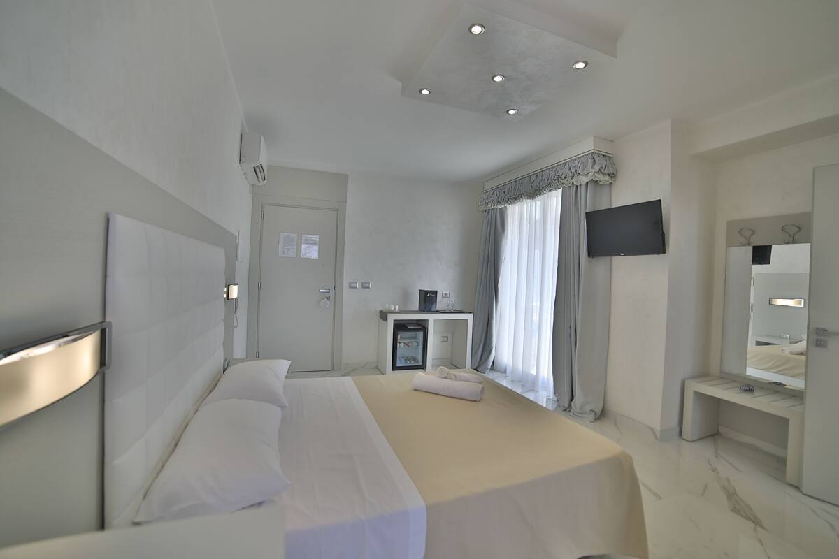 A modern hotel room with a large bed, a bedside table with a lamp, a TV mounted on the wall, and a mirror above the sink.