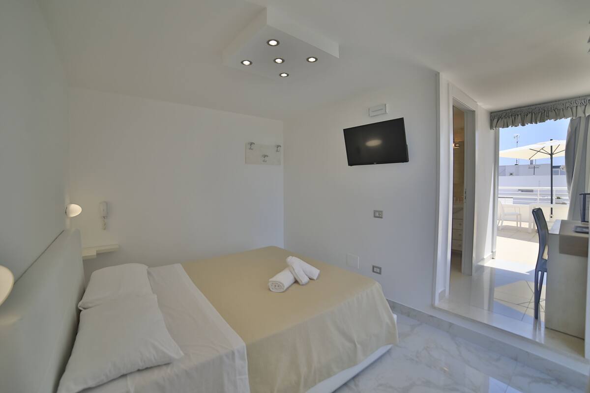 A hotel room with a bed, a flat-screen TV, a window with curtains, and a view of the outside.