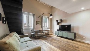 Una sala de estar con un sofá, una mesa de comedor con sillas, un televisor sobre un mueble bajo y una escalera.