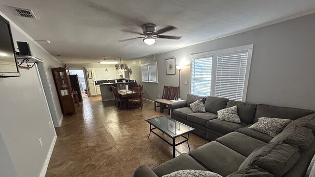 A spacious living room with a gray sectional sofa, a glass coffee table, and a dining area with a kitchen in the background.