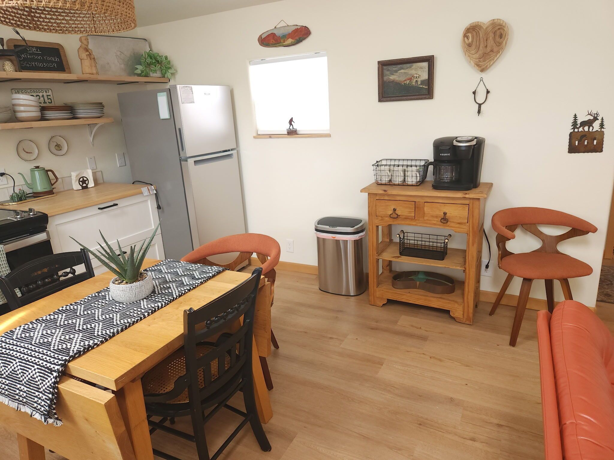 Une cuisine compacte équipée d’un réfrigérateur, d’une cuisinière et d’une machine à café. On y trouve une table à manger avec des chaises et une poubelle.