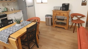 Ett kompakt kök med kylskåp, spis och kaffebryggare. Det finns ett matbord med stolar och en papperskorg.