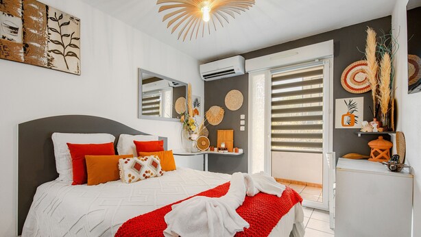 A bedroom with a bed, a bedside table, a mirror, and a window with blinds. - Le Flamboyant du Fonserane - Climatisé (Béziers)