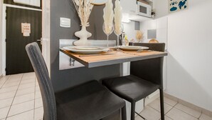 A modern kitchen with a dining table set for two, featuring a vase with dried plants, a microwave, and a shelf with decorative items. - Le Flamboyant du Fonserane - Climatisé (Béziers)
