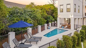A rooftop pool with lounge chairs and an umbrella, surrounded by greenery and a modern building.