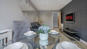 Una sala de estar moderna con una mesa de comedor de vidrio, un sillón tapizado en gris y un sofá. Un televisor de pantalla plana montado en la pared muestra el logotipo de Netflix.