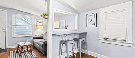 A room with a wooden floor, a grey sofa, a small wooden table, two grey barstools, a white ceiling light, and a window with blinds.