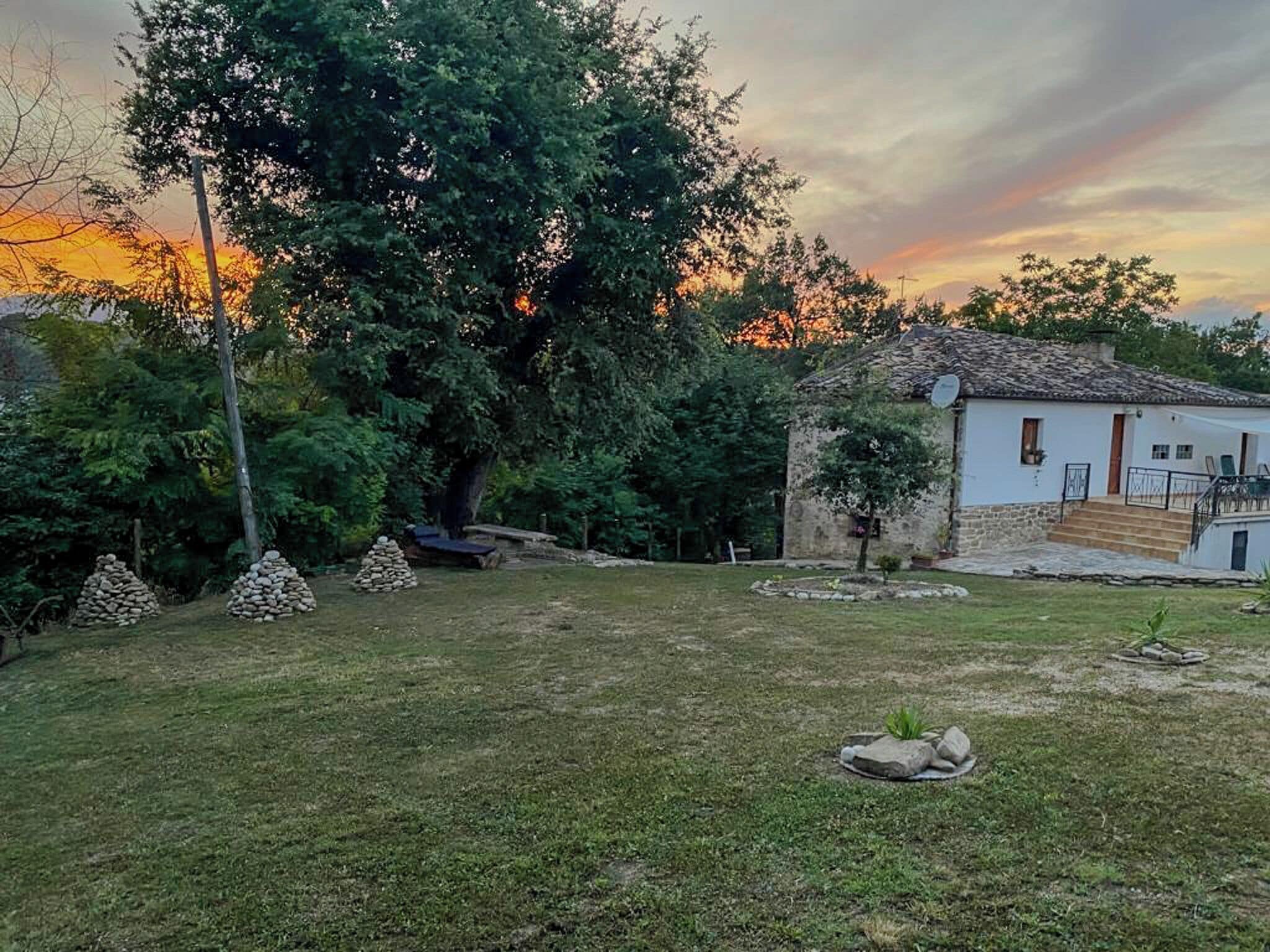 Una casa rústica amb façana de pedra, un jardí petit i un arbre gran al fons.