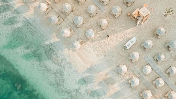 Luftaufnahme eines Strandes mit Liegestühlen, Sonnenschirmen, einem kleinen Boot und einem Rettungsschwimmturm.