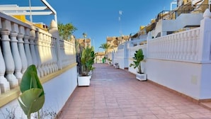 A paved pathway flanked by white walls and balconies, leading to a distant building.