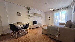 A living room with a sofa, a TV, a dining table with chairs, and a plant on the wall.