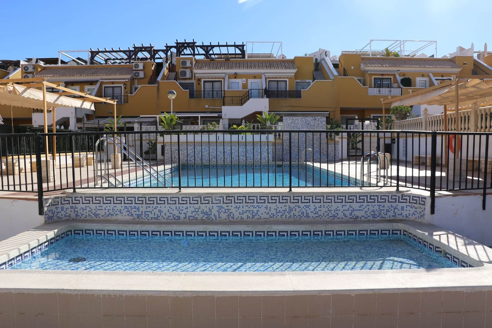 A swimming pool with a tiled border, surrounded by a fence, and adjacent to a building with multiple balconies and a tiled roof.