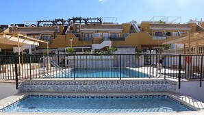 A swimming pool with a tiled border, surrounded by a fence, and adjacent to a building with multiple balconies and a tiled roof.
