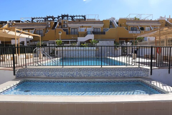 Una piscina con borde de mosaico, rodeada por una cerca, y contigua a un edificio con varios balcones y techo de tejas.
