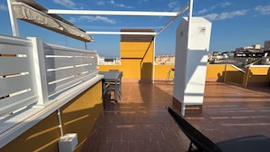 A rooftop terrace with a table and chairs, a white railing, and a view of the cityscape.