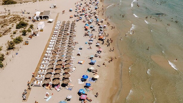 Eine Vogelperspektive zeigt einen Strand mit Reihen von Liegestühlen und Sonnenschirmen, einem Rettungsturm sowie eine sandige Küstenlinie mit Wellen.