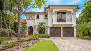 A two-story house with a large front driveway, a garage, and a well-maintained garden with palm trees and shrubs. - Spectacular 4BR/4BA Villa with Private Pool & Parking WiFi, AC (Hacienda Pinilla)