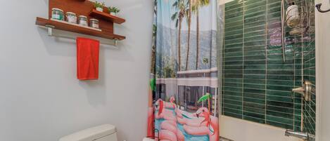 A bathroom with a toilet, a shower with a rain showerhead, a towel rack with a red towel, and shelves with jars and a plant.