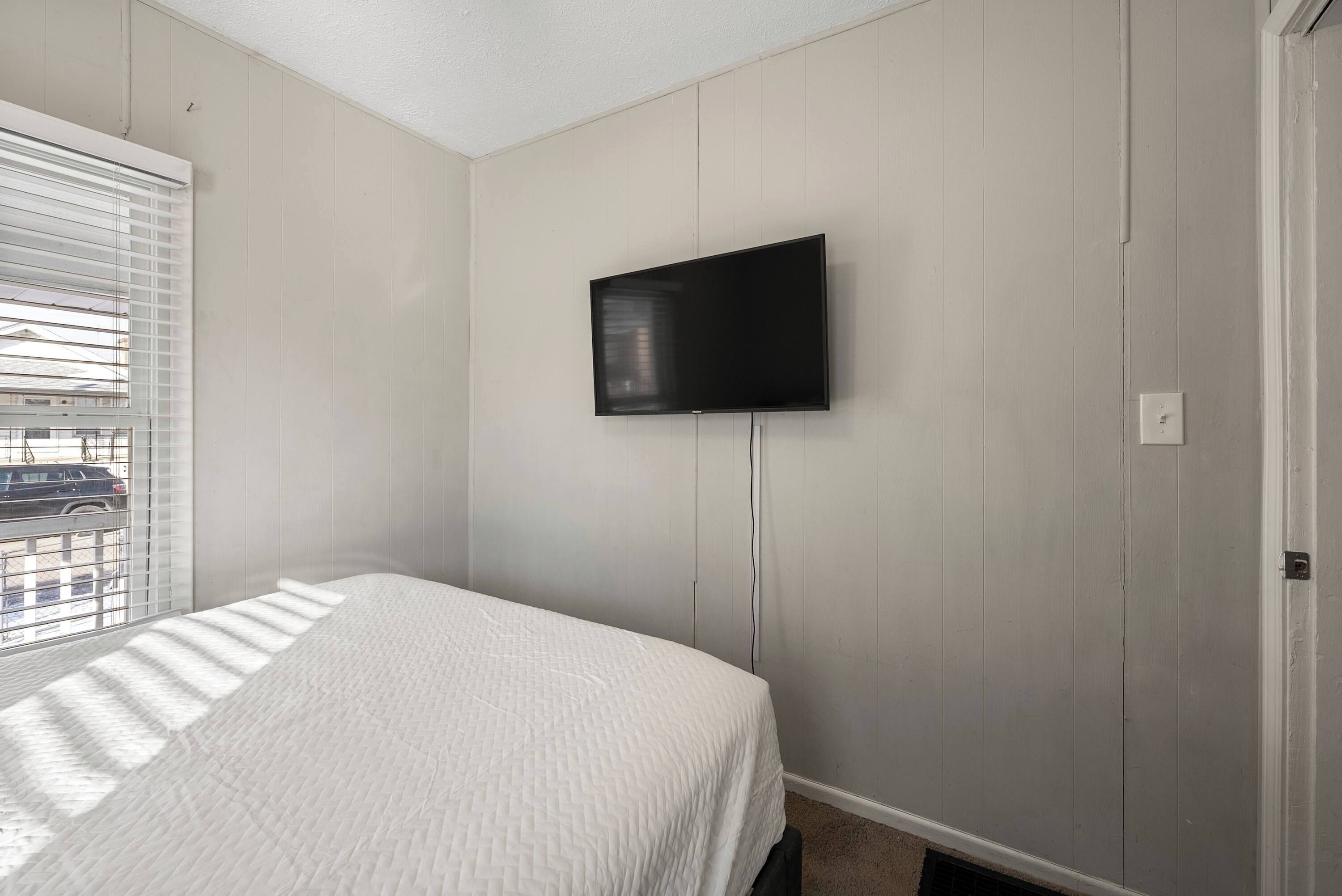 A bedroom with a bed, a television mounted on the wall, and a window with blinds.