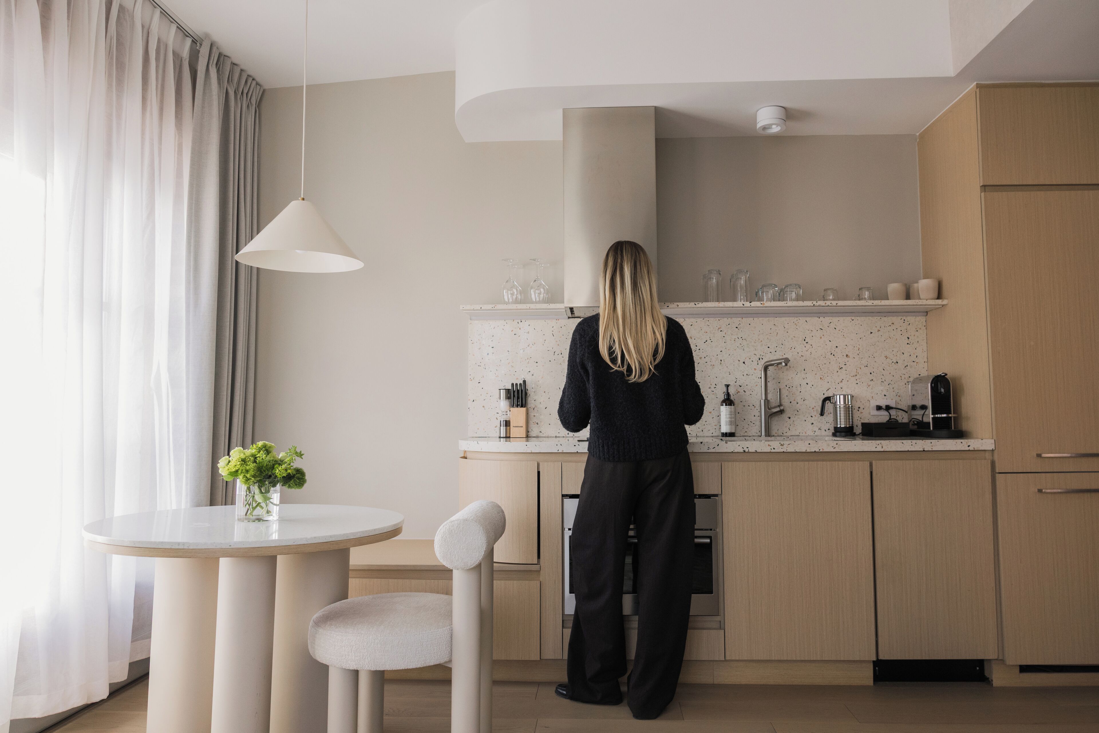 A modern kitchen with a person standing at the counter, a round table with a chair, and a vase with flowers.