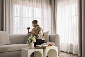 A person sitting on a sofa in a room with a window, a small table with a book and a plate of food, and a vase with flowers. - Corner Apartments  (Montréal)