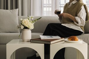 Una persona sentada en un sofá, con un libro, un plato con un croissant y una taza de café sobre una mesita auxiliar.