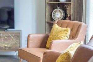 A brown leather armchair with a yellow cushion in a room with a wooden cabinet and a wall-mounted clock. - Lendrick House - 3 bed house with parking, Falkirk (Falkirk)