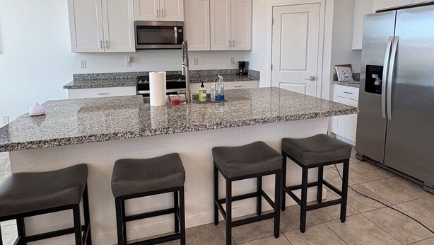 A modern kitchen with white cabinets, a stainless steel refrigerator, a granite countertop, and three bar stools. - Sunlit 4-bedroom West Phoenix Home, Mountain Views & Close to West Gate (Tolleson)