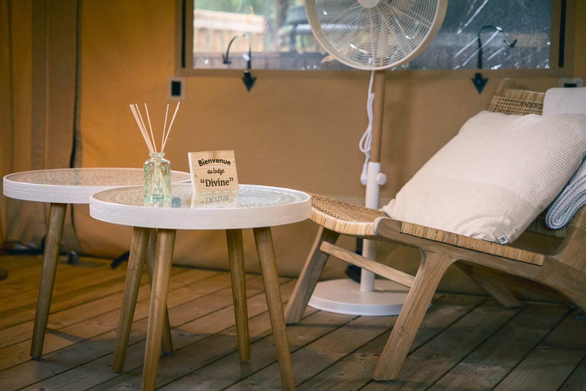 Een houten lounge stoel met een wit kussen, een klein rond tafeltje met een glazen vaas en rietverspreider, en een bordje met de tekst 'Bienvenue au lodge Divine'.