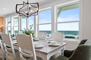 Comedor con vista a la playa, sillas blancas, mesa de madera y una lámpara de araña moderna.