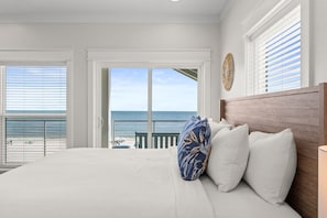 A bedroom with a large bed, white bedding, a wooden headboard, and a view of the ocean through a sliding door.