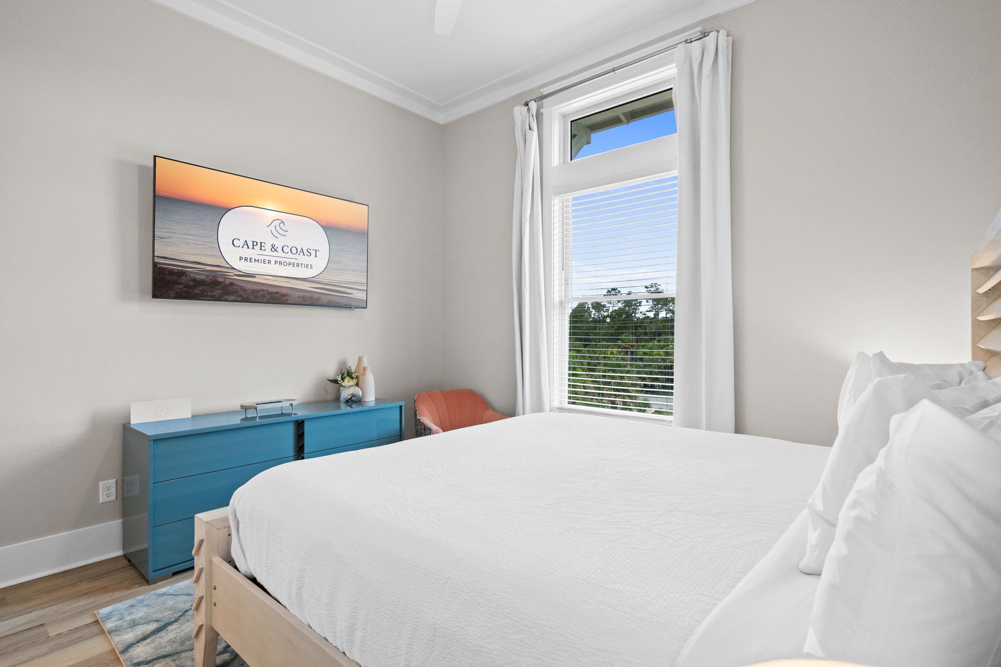Une chambre à coucher avec un grand lit, une commode bleue, un tableau représentant un coucher de soleil et une fenêtre avec des stores.