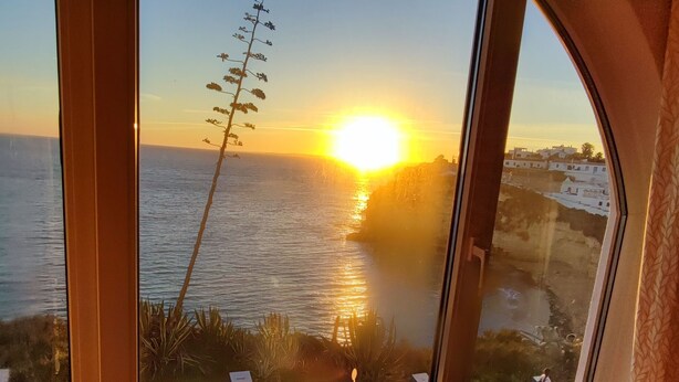 A room with a large window overlooking a coastal sunset, a decorative vase on a table, and a view of a building across the water. - "Ocean View Terrace", The best sea view - directly on the beach of Carvoeiro (Carvoeiro)