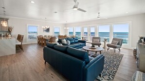 A spacious living room with a large blue sofa, a dining area with wooden chairs, and a view of the ocean through large windows. - Wavy Daze: New Construction, Private Pool, Pet Friendly, Gorgeous Beach Views (St Joe Beach)