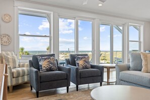 A spacious living room with large windows offering a view of the beach. There are two armchairs with patterned cushions and a sofa with a light-colored throw.