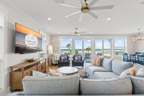 A spacious living room with a large flat-screen TV, a ceiling fan, and a view of the beach through large windows. - Paloma: Beachfront in Mexico Beach, Private Pool, Sleeps 24! (Mexico Beach)