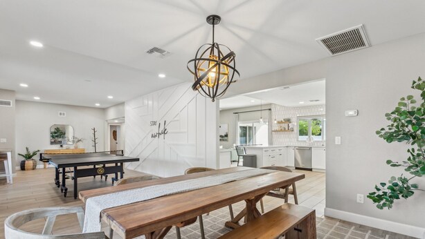 A spacious dining area with a wooden table and benches, a modern kitchen in the background, and a decorative wall with a plant.