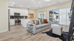 A modern kitchen with white cabinets and stainless steel appliances, a grey sofa with decorative pillows, and a large sliding glass door leading to an outdoor area.