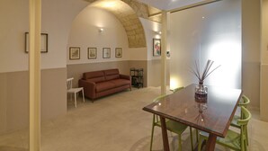 A modern living room with a brown sofa, a glass dining table, and green chairs. There is a stone archway and a wall with framed pictures.