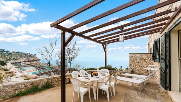 A covered outdoor dining area with a table and chairs, overlooking a scenic coastal view.