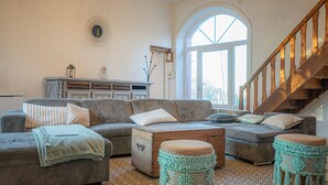 A cozy living room with a large window, a wooden staircase, and a decorative wall clock. - Le Florentin - Spacious Family Home in a Peaceful Village near Limoges (Condat-sur-Vienne)