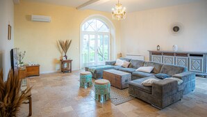 A spacious living room with a large sectional sofa, a chandelier, and a view through a large arched window. - Le Florentin - Spacious Family Home in a Peaceful Village near Limoges (Condat-sur-Vienne)