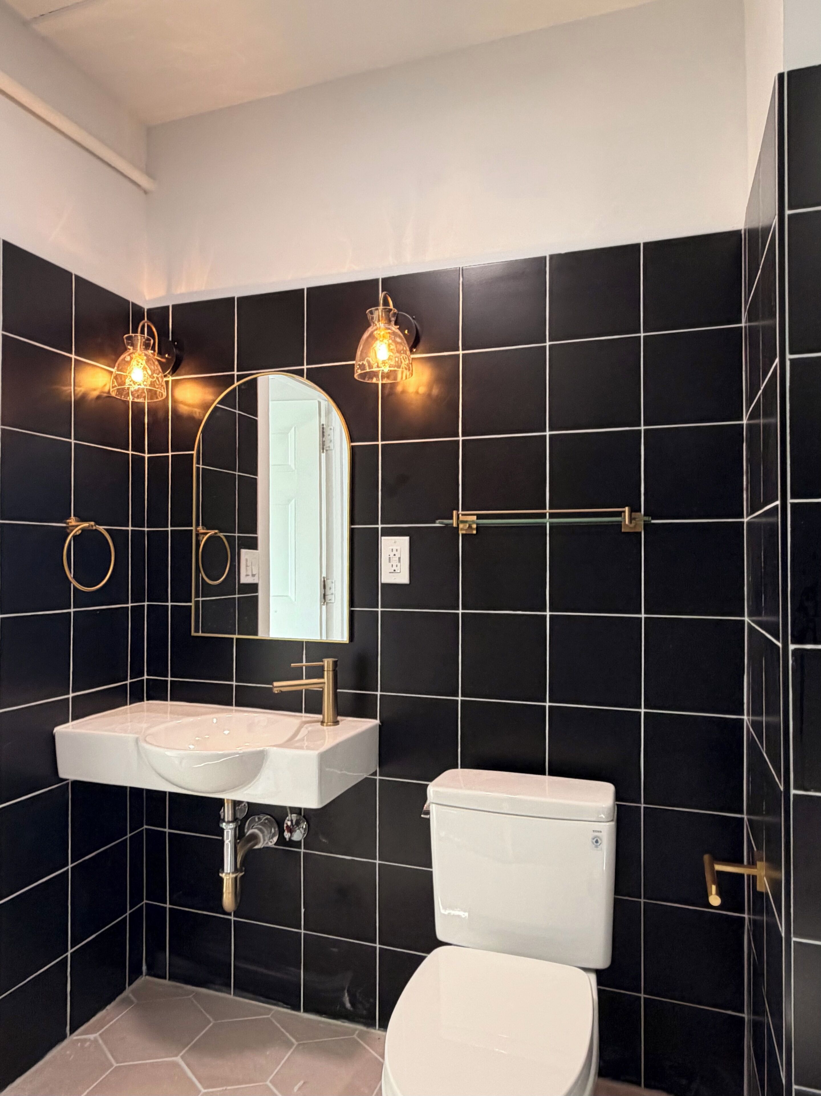 A modern bathroom with black tiled walls, a white sink, a toilet, and two wall-mounted light fixtures.