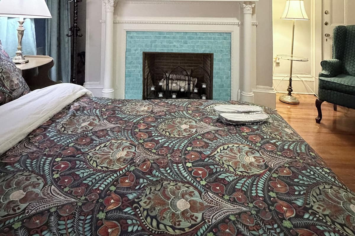 A bedroom with a patterned bedspread, a fireplace, a ceiling fan, and a green armchair.