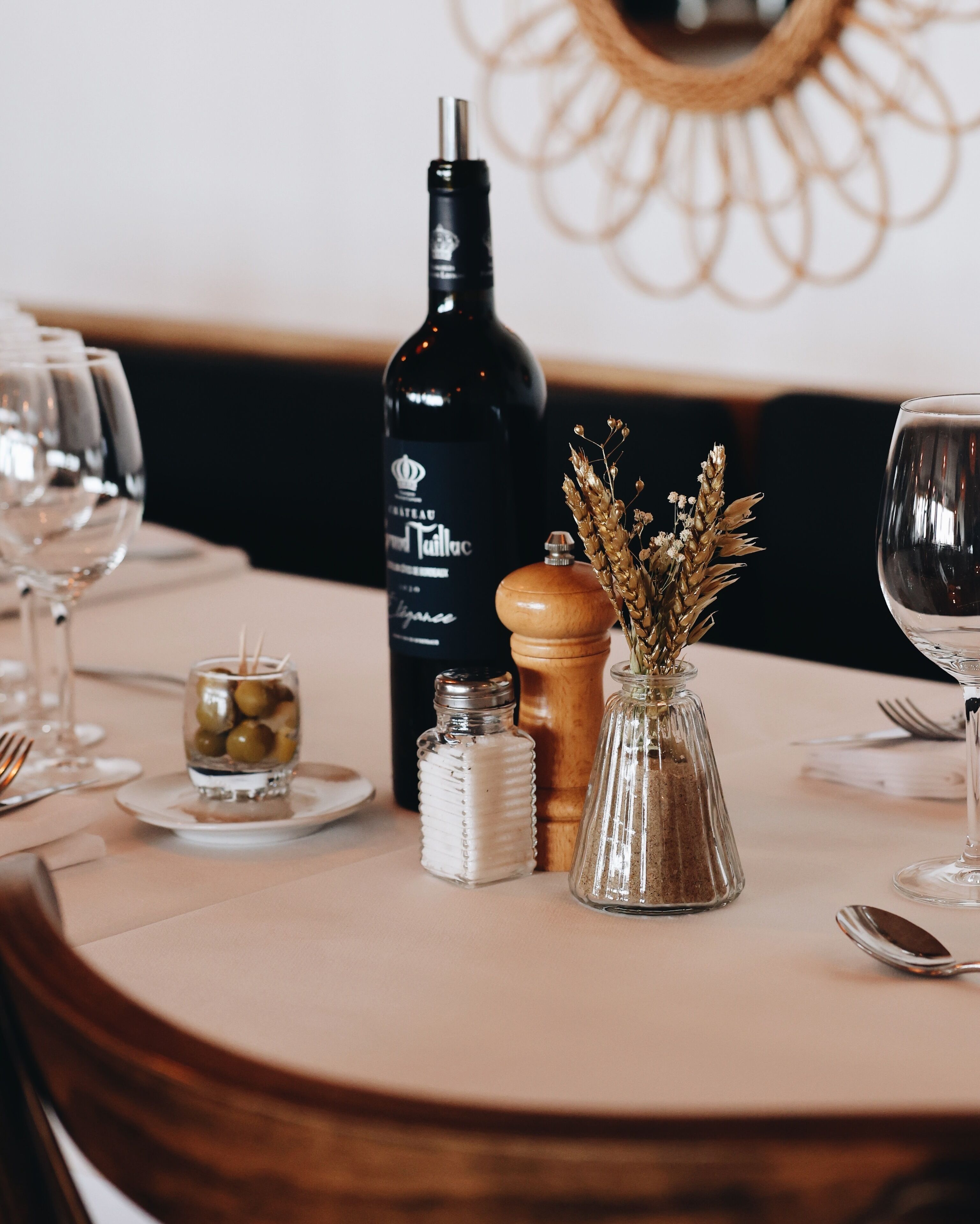 Une table à manger dressée avec une bouteille de vin de Château Moutaut, un salier et un poivrier, un petit verre contenant des olives et un vase avec des fleurs séchées.