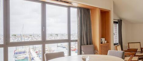 Amplia habitación con una gran mesa redonda y seis sillas, ventanas grandes con vistas a barcos y un techo inclinado.