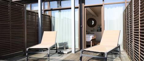 Un espacio al aire libre iluminado por el sol con dos sillones de descanso, una terraza de madera y un ventanal que da a una habitación con una cama y un espejo redondo.