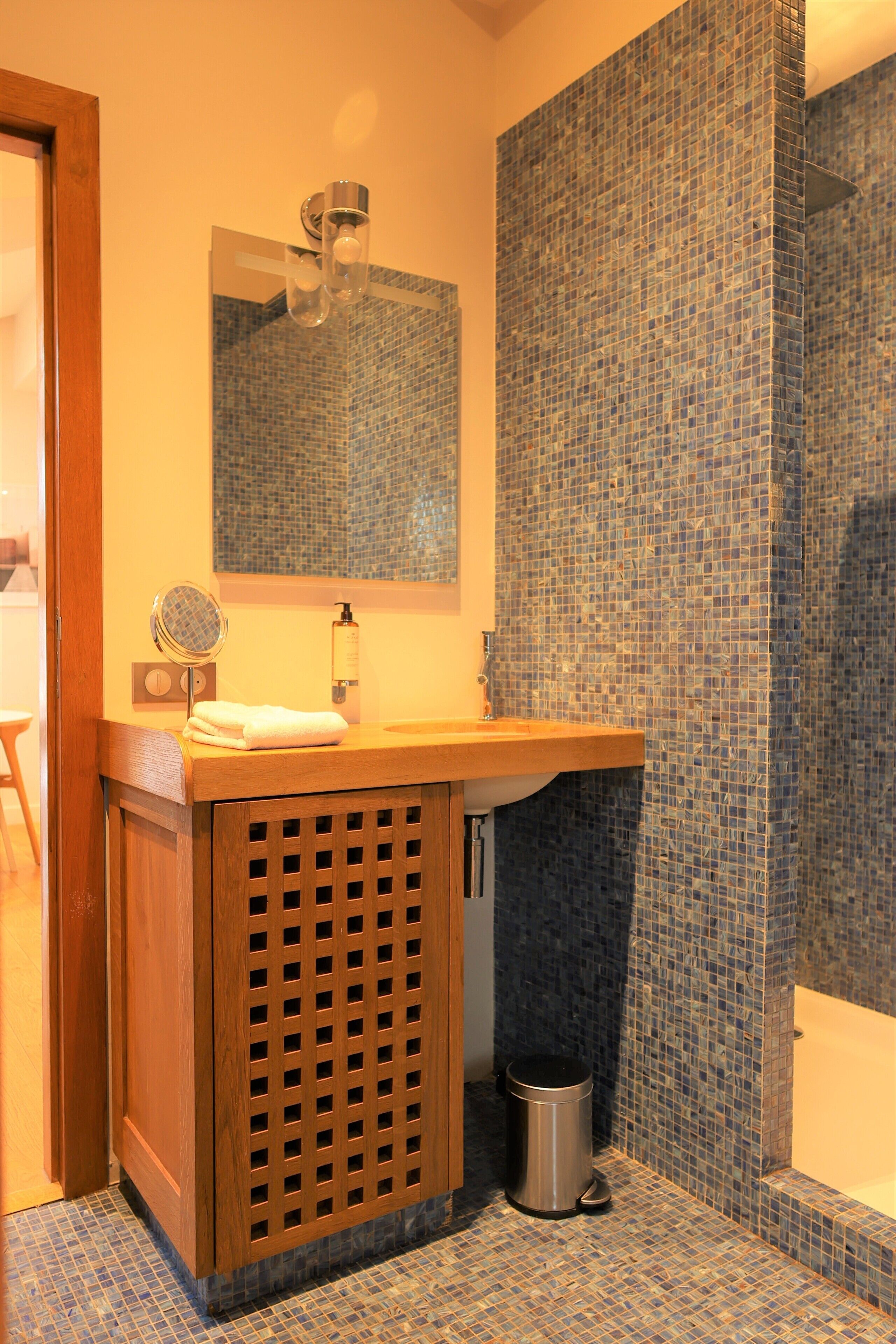 Ein Badezimmer mit einem Holzbadezimmerhocker, einem Spiegel, einem Waschbecken und einem gefliesten Duschbereich.