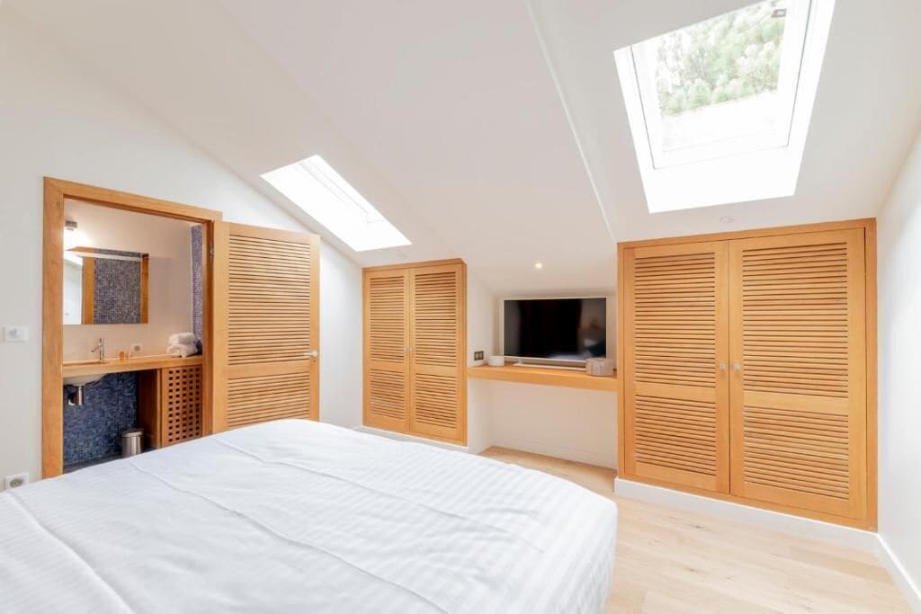 Ein Schlafzimmer mit einem Bett, Dachfenstern, einem Fernseher, Holzschränken und einem Badezimmer mit Waschbecken und Spiegel.